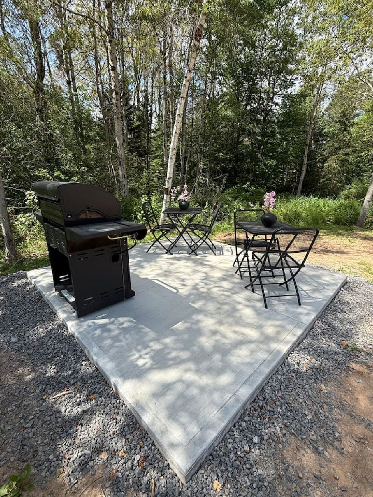 Un espace barbecue extérieur avec une table et des chaises, entouré de gravier et d’arbres.