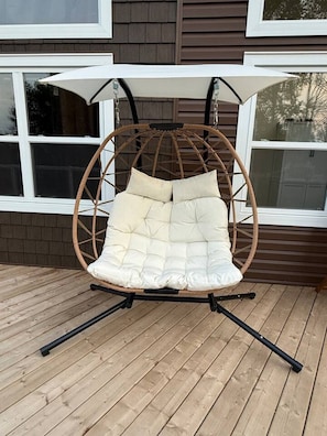 Una poltrona a dondolo in vimini con seduta e schienale imbottiti, sostenuta da una struttura in metallo, su un pontile in legno.