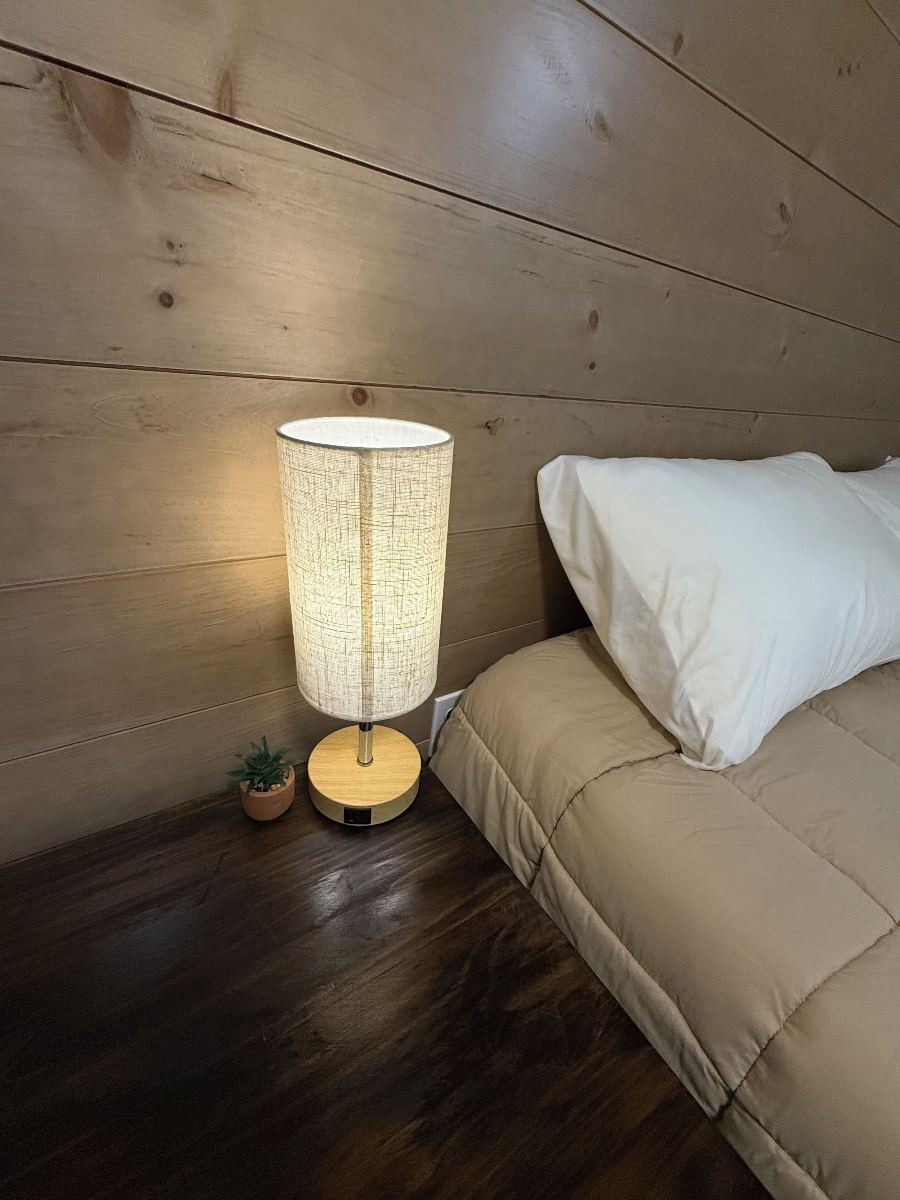 Uma mesa de cabeceira de madeira com uma luminária, uma pequena planta em vaso, e uma cama com roupa de cama branca.
