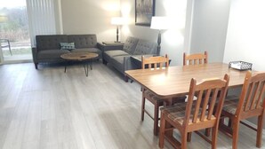 Ein Wohnzimmer mit einer Couch, zwei Beistelltischen mit Lampen, einem Holzessttisch mit Stühlen und einem großen Fenster mit Jalousien.
