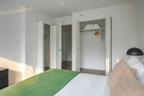 Ein Schlafzimmer mit einem Bett, einer grünen Strukturmatte, einem weißen Kopfkissen, einer schwarzen Lampe und einem Kleiderschrank mit gefalteten Handtüchern.