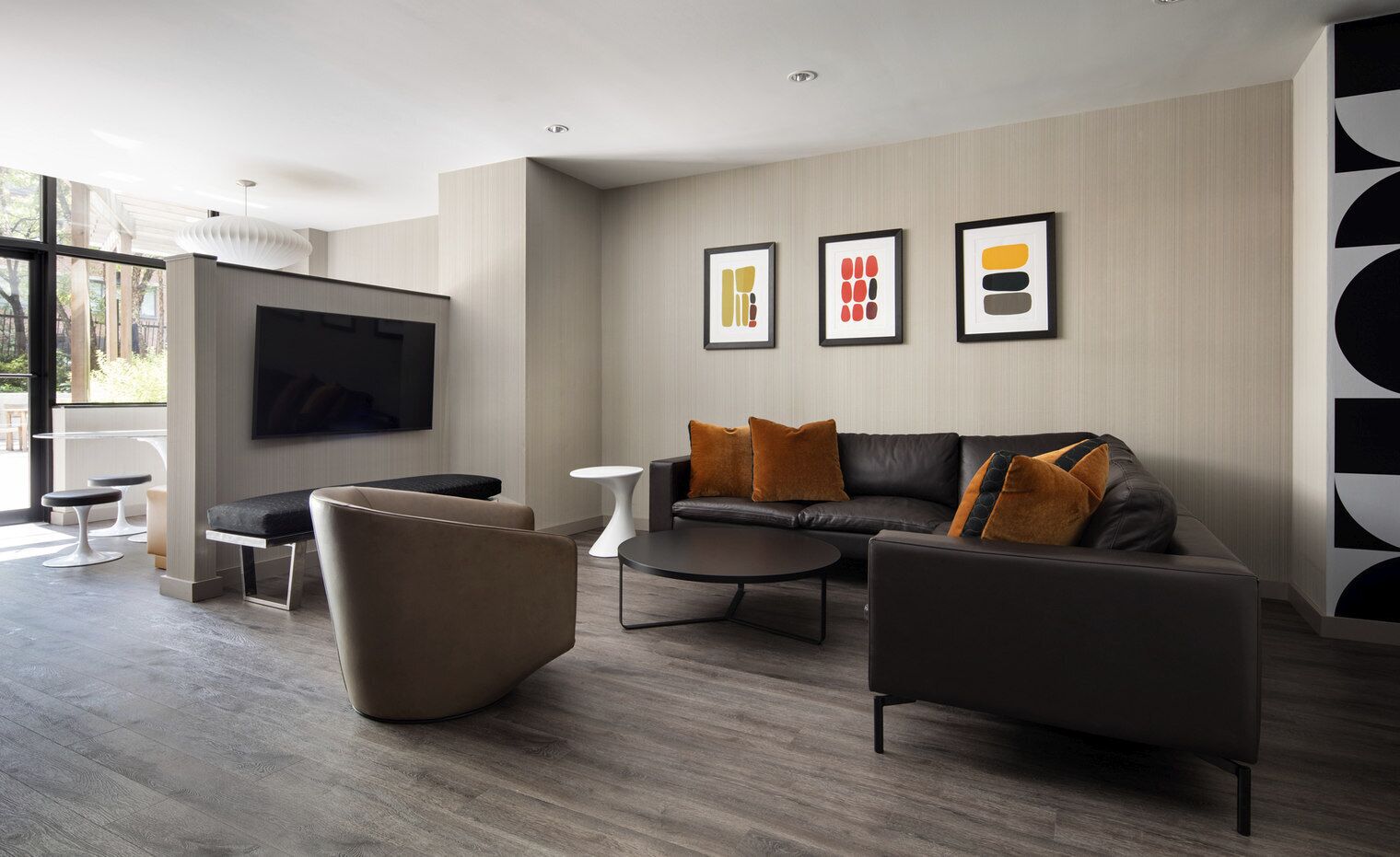 A modern living room with a sofa, armchairs, a coffee table, and a TV. There are framed artworks on the wall and a large window providing natural light.