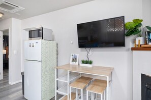 A compact living space with a kitchenette, a flat-screen TV mounted on the wall, a small wooden table, and a potted plant. - Avocado Hut- HOT TUB, free parking (San Diego)