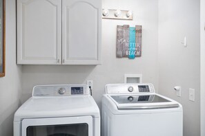 A laundry room with two white front-loading washers and dryers, a wall-mounted light fixture, and a decorative sign that reads 'Life is BETTER at the BEACH.' - Avocado Hut- HOT TUB, free parking (San Diego)