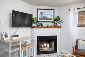 A living room with a flat-screen TV mounted on the wall, a fireplace with a mantel, and a wooden table with a white metal frame. - Avocado Hut- HOT TUB, free parking (San Diego)