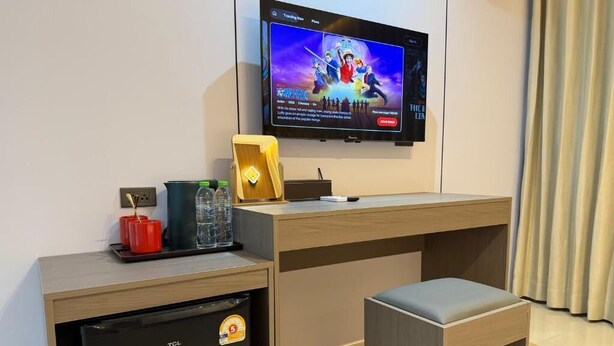 Ett hotellrum med en tv som är monterad på väggen, ett litet kylskåp, ett skrivbord i trä, en vadderad bänk och ett bricka med en röd mugg och glas.