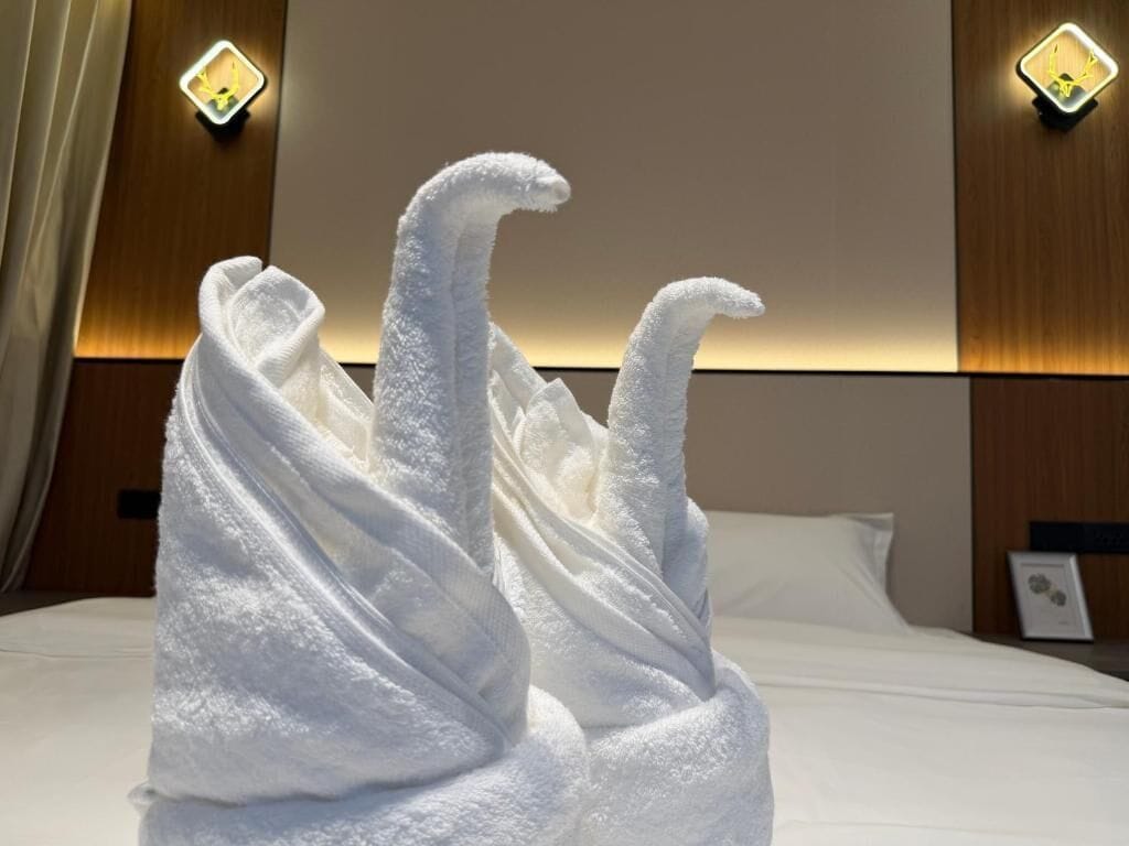 A neatly made bed with a white towel arranged in a decorative manner, a wooden headboard, and wall-mounted light fixtures.