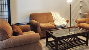 Een woonkamer met twee bruine banken, een glazen salontafel, een vloerlamp en een witte muur met een geometrisch schap.