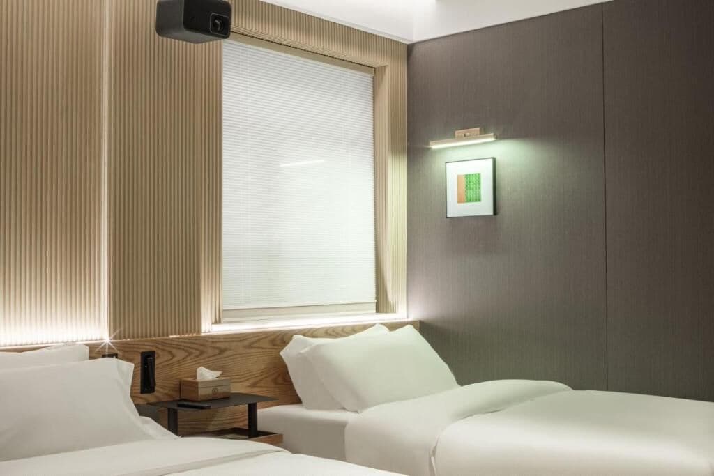 A hotel room with a bed, bedside table, and a window with blinds.