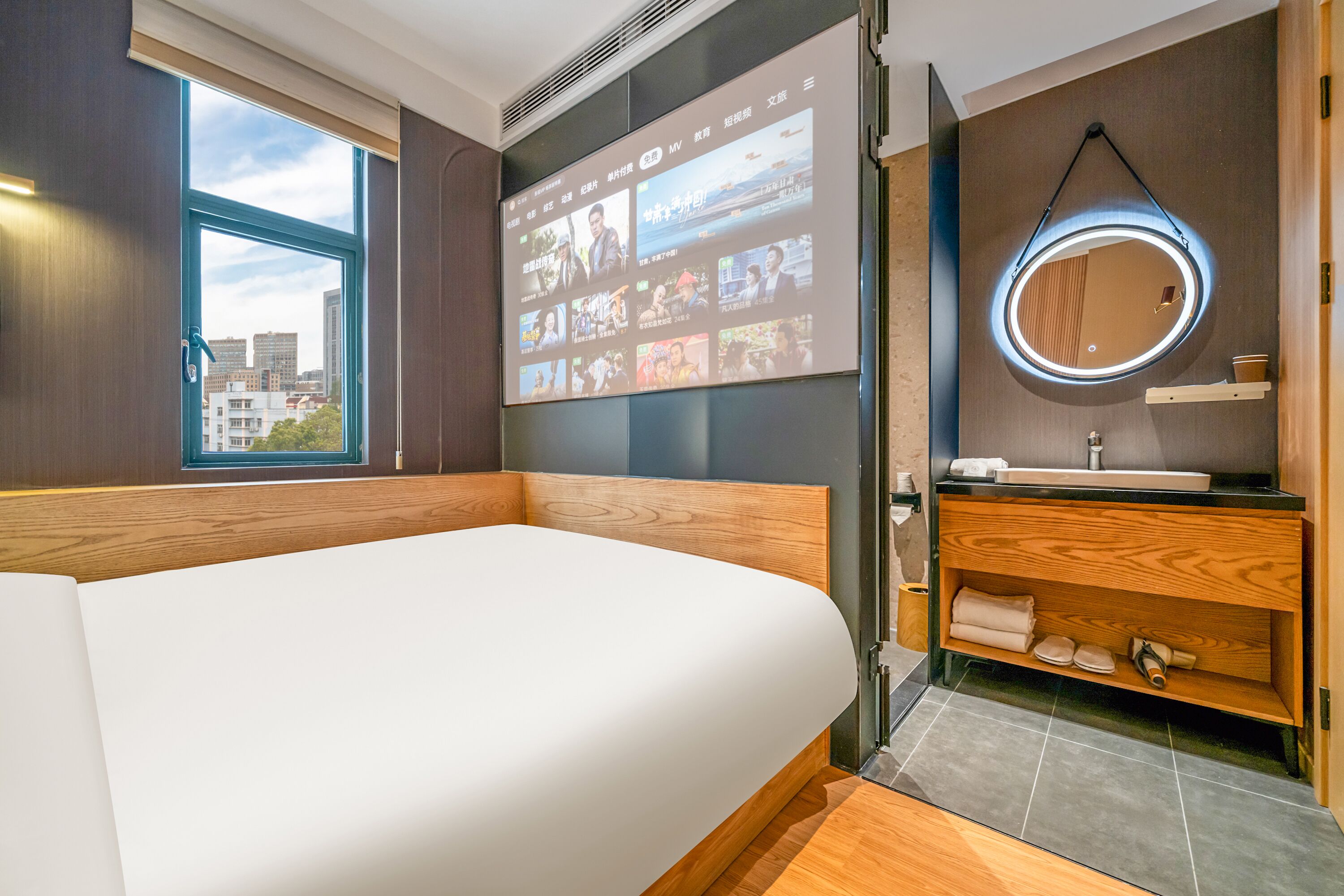 Quarto de hotel moderno com cama, TV fixada na parede exibindo diversos programas, espelho redondo e vista da cidade pela janela.