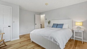 Ein Schlafzimmer mit einem weißen Bett, einem Nachttisch mit Lampe, Holzboden und einer Tür.