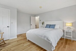 Ein Schlafzimmer mit einem weißen Bett, einem Nachttisch mit Lampe, Holzboden und einer Tür.
