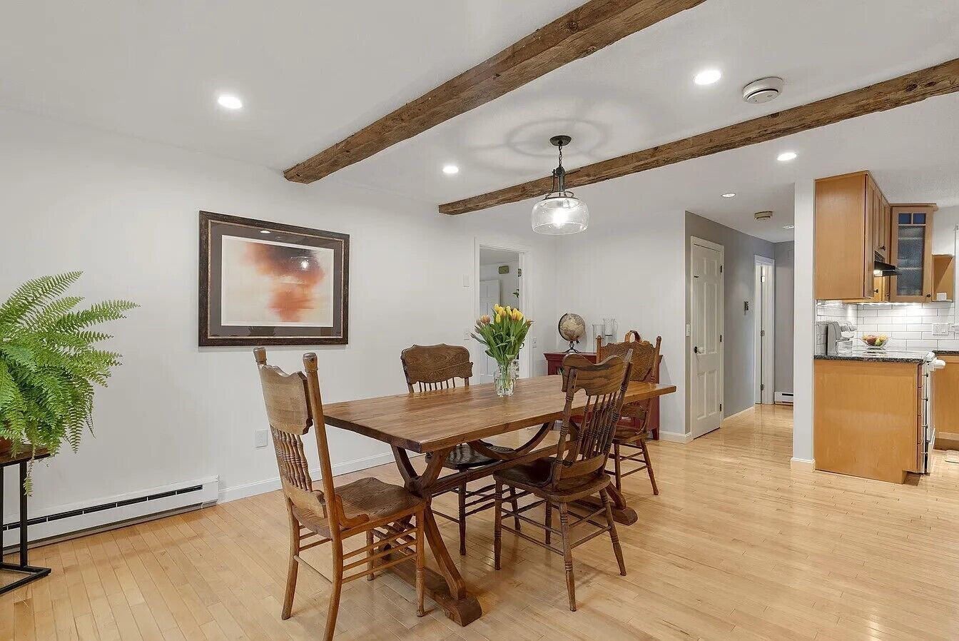 A dining area with a wooden table and chairs, a ceiling with exposed beams, and a kitchen in the background.