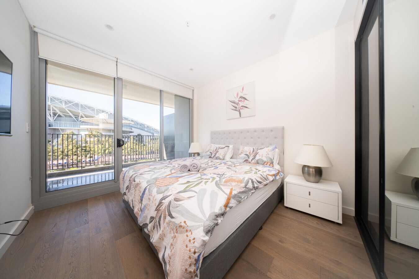 A bedroom with a large bed, a nightstand, a lamp, and a view of a building through a glass door.