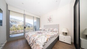 Ein Schlafzimmer mit einem großen Bett, einem Nachttisch, einer Lampe und Blick auf ein Gebäude durch eine Glastür.