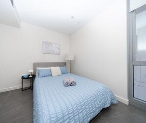 A bedroom with a bed, a nightstand, a lamp, and a window. - Modern Retreat near stadiums (Lidcombe)