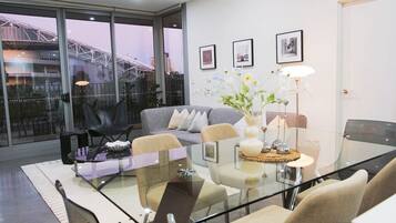A modern living room with a glass table, grey sofa, and a view of a bridge through large windows.