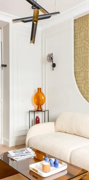 A modern living room with a sofa, a coffee table with magazines and a tray, a wall-mounted light fixture, and a patterned wallpaper. - PREMIUM 3-BEDROOM APARTMENT IN A PRESTIGIOUS PARIS NEIGHBORHOOD (Paris)