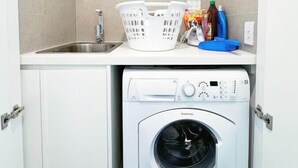 A compact laundry area with a washing machine, a sink, and a countertop with laundry baskets and cleaning supplies. - Sydney Olympic Park View Bliss | Modern Design (Sydney Olympic Park)