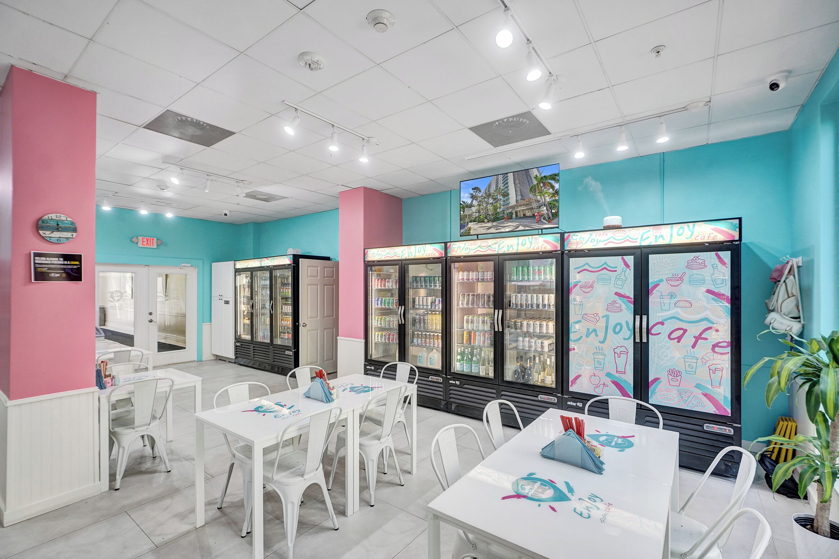 A cafe with white tables and chairs, teal walls, and a large refrigerator with colorful drink options.