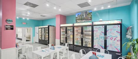 A cafe with white tables and chairs, teal walls, and a large refrigerator with colorful drink options.