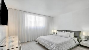 A hotel room with a bed, two bedside tables with lamps, a TV, and a large window with sheer curtains. - Haven Beach Gem Condo in Hollywood (Hollywood)