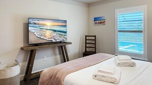 Une chambre à coucher avec un lit, un téléviseur posé sur un meuble, une fenêtre avec des stores et un tableau accroché au mur.