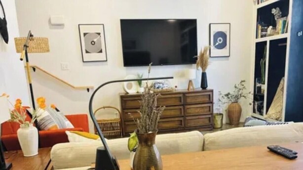 A living room with a wooden coffee table, a vase with dried plants, a sofa, a television mounted on the wall, and a bookshelf.