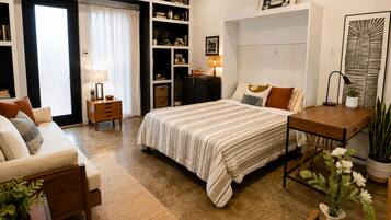 A bedroom with a bed, bedside table, lamp, and a desk with a chair. There is a bookshelf with various items and a window with curtains.