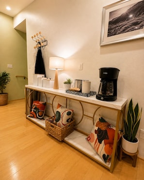 A modern living room with a wooden floor, a white console table, a coffee maker, a lamp, and decorative pillows.