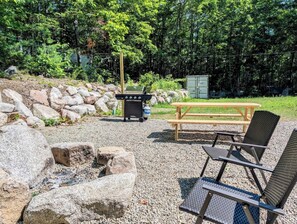 Zona de pícnic amb una barbacoa, taula i cadires, envoltada de roques i arbres.