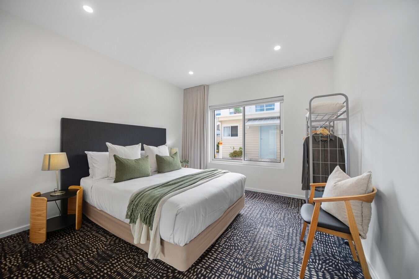 A hotel room with a bed, a bedside table with a lamp, a window with curtains, a metal rack with clothes, and a patterned carpet.