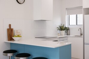 A modern kitchen with a white and blue color scheme, a built-in island with bar stools, a clock on the wall, and a window providing natural light. - Esplanade Apartments | Ocean View Beachfront | West Beach (West Beach)