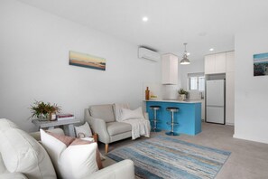 A modern living room with a sofa, a coffee table, and a rug. There is a kitchen area with a blue countertop and a refrigerator. - Esplanade Apartments | Ocean View Beachfront | West Beach (West Beach)