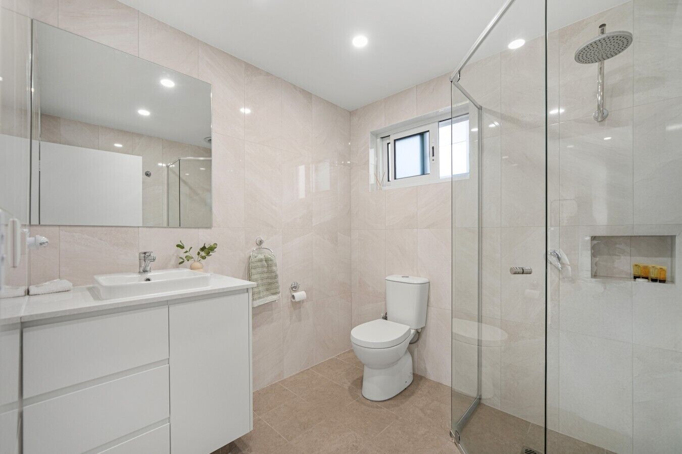 A modern bathroom with a glass shower enclosure, a toilet, a sink with a mirror, and a small window above the toilet.