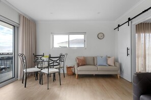 A modern living room with a sofa, dining table, chairs, and a large window. - Esplanade Apartments | Ocean View Beachfront | West Beach (West Beach)