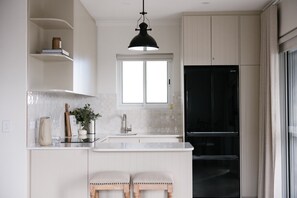 Une cuisine moderne avec des meubles blancs, un luminaire suspendu noir, une fenêtre au-dessus de l’évier et un réfrigérateur noir.