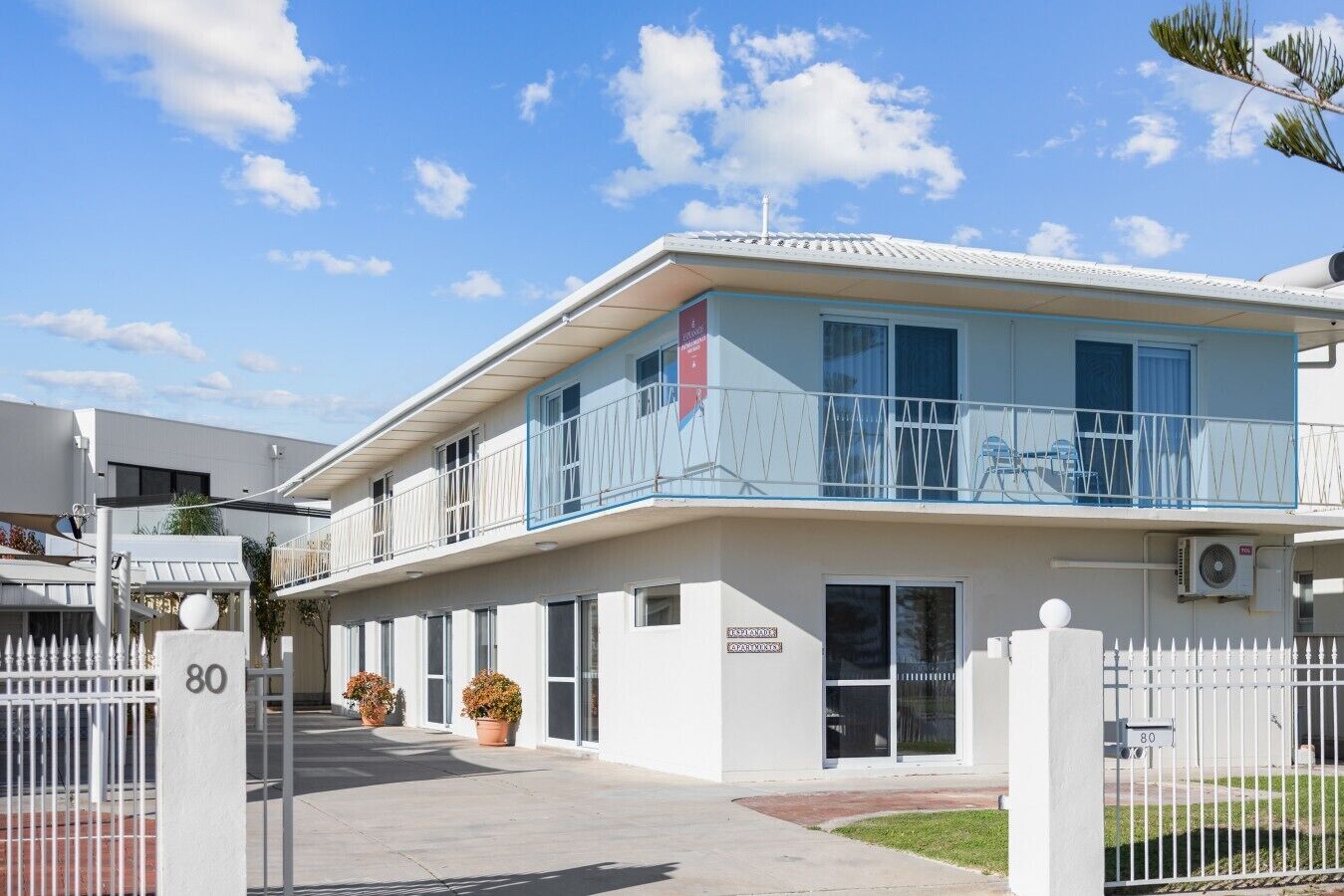 A modern building with a white facade, a balcony with a blue railing, and a gated entrance.