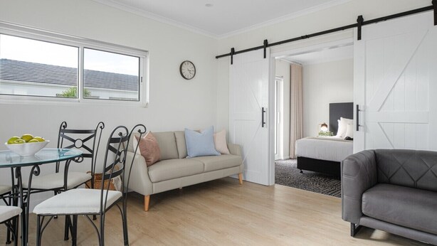 A modern living room with a dining area, a sofa, a grey armchair, a bed, and a sliding barn door. - Esplanade Apartments | Ocean View Beachfront | West Beach (West Beach)
