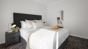A modern bedroom with a large bed, a nightstand with a lamp, and a mirror. - Esplanade Apartments | Ocean View Beachfront | West Beach (West Beach)