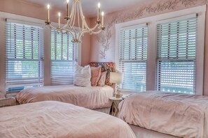 A bedroom with two beds, a chandelier, and large windows with blinds.