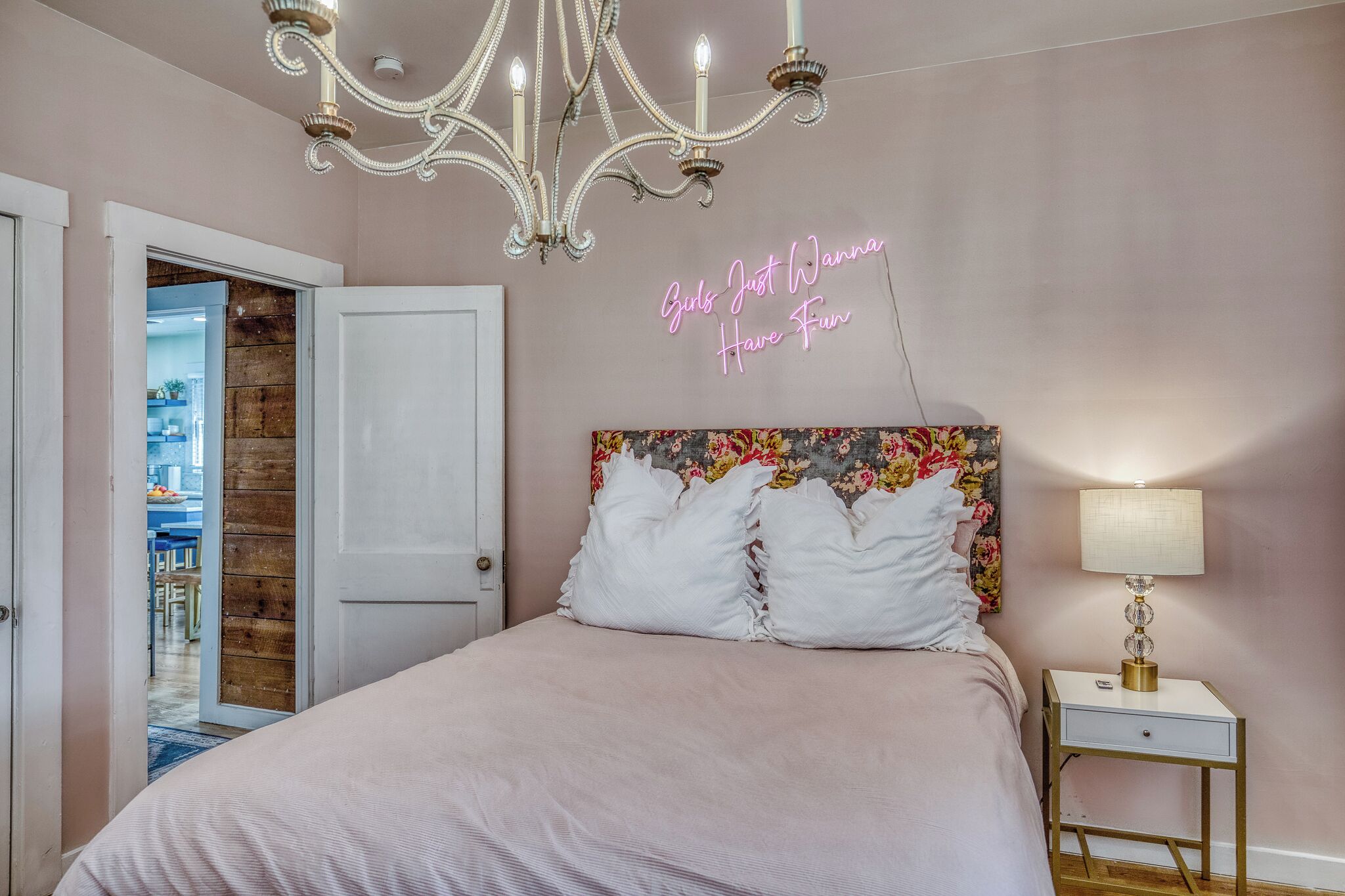 A bedroom with a chandelier, a neon sign, a floral headboard, and a bedside table with a lamp.