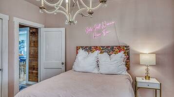 A bedroom with a chandelier, a neon sign, a floral headboard, and a bedside table with a lamp.