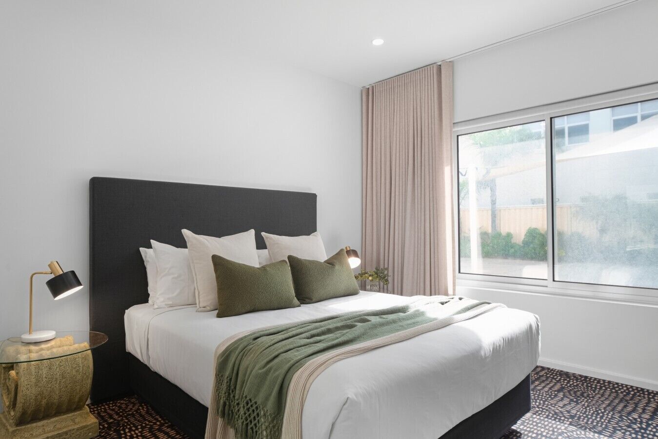 A modern hotel room with a large bed, a bedside lamp, and a view of the outdoors through a large window.