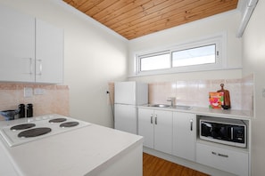 A compact kitchen with white cabinets, a stove, a refrigerator, a microwave, and a window above the sink. - Esplanade Apartments | Ocean View Beachfront | West Beach (West Beach)