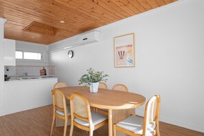 A dining area with a wooden table and chairs, a potted plant, a clock, and a kitchen in the background. - Esplanade Apartments | Ocean View Beachfront | West Beach (West Beach)
