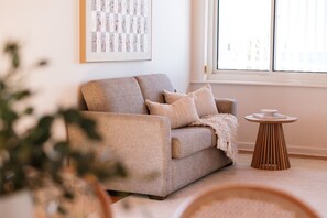 A neutral-toned sofa with cushions, a small wooden side table, and a framed abstract artwork on the wall. - Esplanade Apartments | Ocean View Beachfront | West Beach (West Beach)