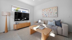 Ett modernt vardagsrum med en platt-TV, ett träskåp, en beige soffa med kuddar, ett soffbord och ett stort fönster.