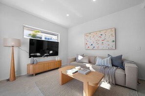 Ein modernes Wohnzimmer mit Flachbildfernseher, einer Holzkabine, einem beigefarbenen Sofa mit Kissen, einem Couchtisch und einem großen Fenster.