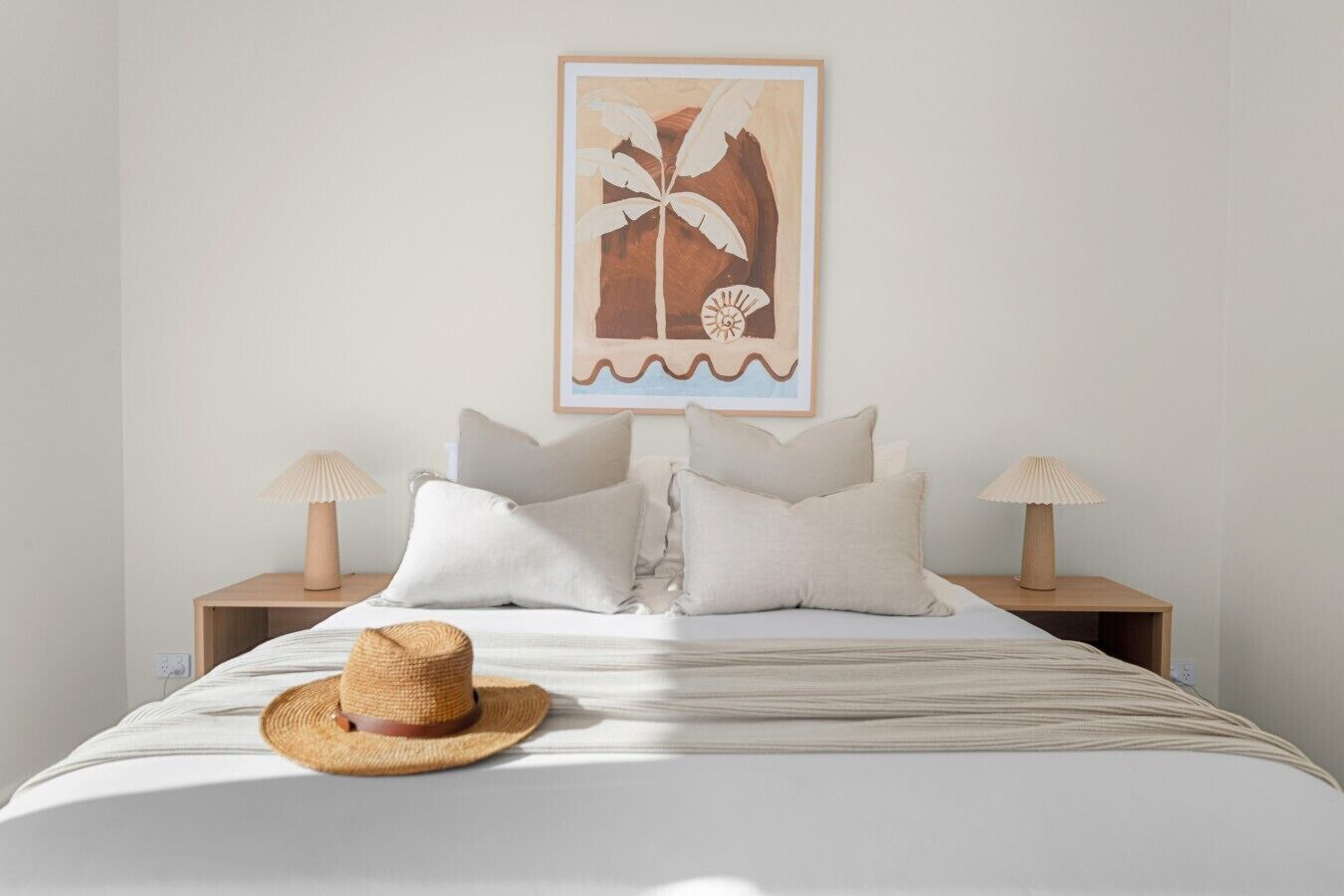 A bed with a straw hat, two bedside tables with lamps, and a framed picture on the wall.